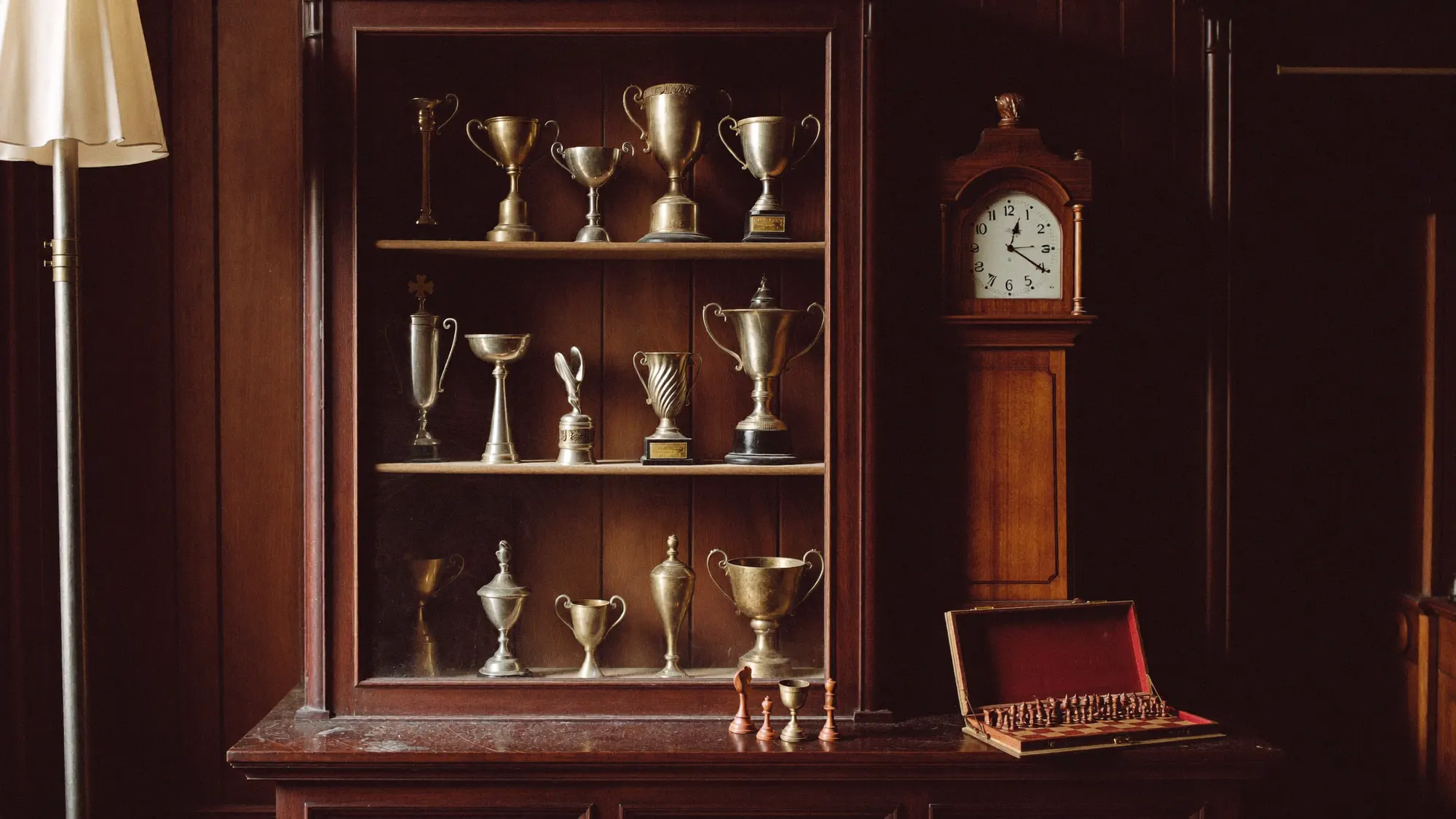 Une vitrine ancienne en bois sombre garnie de coupes en laiton ternies, à côté d'une horloge de parquet